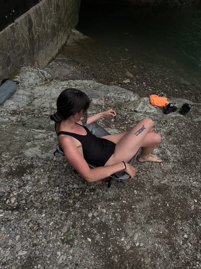 A person with dark hair pulled back in a bun, wearing a black one-piece swimsuit, sits in a gray camping chair on a rocky shore. Their right leg is extended, revealing a black tattoo on their thigh. An orange towel and a pair of black sandals are on the ground to their right. A gray bag is visible to the left of the chair. The person is looking down at something in their hand. The background shows a stone wall and dark water. The scene appears to be an outdoor natural setting, possibly near a river or lake. The image does not provide any specific information about the location in the city of Khleborob, Russia.