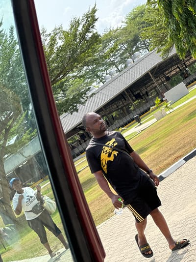 The image depicts an outdoor scene in Ozuaha, Nigeria, under bright daylight. The perspective is from inside a vehicle, indicated by a dark-colored structural element and partial reflections on the left side of the frame.
In the foreground, slightly to the right, a dark-skinned adult male stands on a light-colored, patterned paved surface. He wears a black short-sleeved t-shirt featuring a gold or yellow stylized lion graphic, black shorts with three gold or yellow stripes on the left leg, and black slide sandals with "BOTTEGA" in gold/yellow text. A black shoulder bag is worn across his chest. He holds a clear plastic water bottle with a light green cap in his left hand. His gaze is directed towards the viewer's left.
Further back and to the left, partially obscured by reflections, a dark-skinned adult female stands on a grassy area. She wears a blue baseball cap, a white short-sleeved t-shirt with a dark logo, dark capri pants, and light-colored sandals. She has a black fanny pack or waist bag and carries a light-colored bag over her left shoulder. Her right arm is raised.
The background features a maintained grassy area and numerous green trees. A large, open-sided structure with a light-colored, sloped roof is visible behind the grass. The roof is equipped with multiple dome-shaped ventilation units. Rows of seating are discernible beneath the structure. A black and white striped curb or barrier separates the paved surface from the grassy area. The sky above is light blue with sparse white clouds.