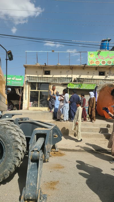 The image displays a sunny outdoor street scene in Kuchlak, Pakistan, with a clear blue sky and scattered white clouds. In the foreground on the right, a large piece of heavy machinery, possibly a front-end loader, is visible, showing its robust tire and part of its hydraulic arm. Strong sunlight casts distinct shadows of people and objects onto the concrete ground, which has scattered dirt and sand.

On the left side of the street, a row of buildings lines the sidewalk. Several men, dressed in traditional shalwar kameez attire, are gathered on the sidewalk, standing casually, some appearing to observe their surroundings or interact. One man near the center is looking at a mobile phone.

Multiple signs are affixed to the buildings. A prominent green banner sign displays text in an Arabic script, reading "رحمت البتول" (Rahmat-ul-Batool), alongside illustrations of various food items including tea, and a partial phone number "0315-832". Below, another green sign bears the name "عبداللہ" (Abdullah) and a full phone number "0336-9732338". A blue water tank is visible on the roof of one building, and overhead power lines crisscross the upper portion of the sky. An orange tarp or fabric covers an object in the background near the buildings.