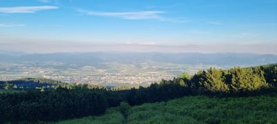 This is a panoramic, wide-angle photograph taken from a high vantage point, likely a mountain overlooking the city of Martin, Slovakia. The foreground is dominated by lush green vegetation, including low-lying bushes and the tops of coniferous trees, suggesting a natural, possibly mountainous, environment. The middle ground reveals the urban sprawl of Martin, characterized by dense clusters of buildings with red-tiled roofs interspersed with lighter-colored structures. The city is nestled within a valley, surrounded by rolling hills and distant mountain ranges that fade into a hazy blue due to atmospheric perspective. The sky is a clear, bright blue, with a few wispy white clouds scattered across it, indicating fair weather. The lighting suggests it is either early morning or late afternoon, with the sun casting long shadows and illuminating the landscape with a warm glow. The overall scene conveys a sense of tranquility and a vast expanse of natural and urban landscapes.
