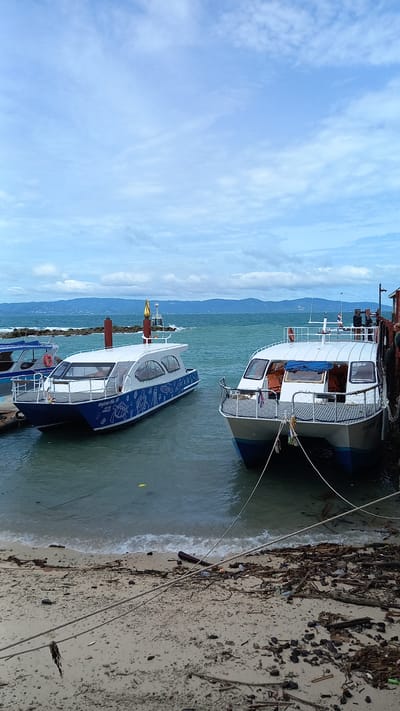 Two white and blue passenger catamarans are docked at a sandy beach in Ban Haad Rin, Thailand. The boat on the left is decorated with white illustrations of marine life. The boat on the right is tied to a wooden pier with thick ropes. The water is calm and a few waves are breaking on the shore. The beach is littered with debris, including driftwood, plastic bottles, and fishing nets. In the background, a range of hills is visible under a cloudy sky.