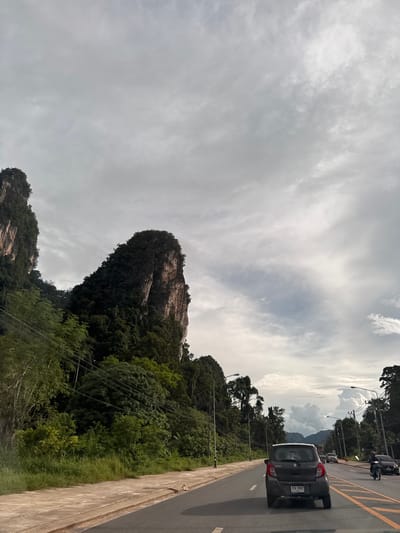 The image shows a road in Ao Luek, Thailand, with a gray Suzuki Celerio car in the foreground, driving on the right side of the road. The car has a license plate that reads "กผ 7867". To the left of the road, there is a concrete sidewalk and a lush green embankment covered in trees and vegetation, with power lines strung across it. In the background, a large, steep limestone karst mountain, covered in greenery, dominates the landscape. The sky is overcast with scattered clouds. Other vehicles, including a motorcycle and several cars, are visible further down the road. The scene appears to be a typical roadside view in a tropical environment.
