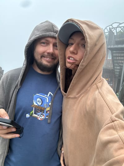 Two people are posing for a selfie. The person in the foreground is wearing a gray hooded sweatshirt and a blue t-shirt with a cartoon design. They are holding a black object in their hand. The person in the background is wearing a brown hooded sweatshirt and a gray baseball cap. They appear to be outside in a foggy environment, with what seems to be a tall structure visible in the upper background. The location might be Binh Lu, Vietnam, based on the structure in the background.