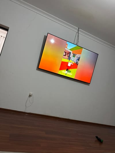 The image shows a living room with a television mounted on the wall. The television is displaying a video game, possibly Minecraft. The room has light-colored walls and a wooden floor. There is a white electrical outlet on the wall with a charger plugged in. The ceiling has a water stain. The overall scene is unremarkable and does not provide any specific information about the location being in Serebryakovka, Russia.