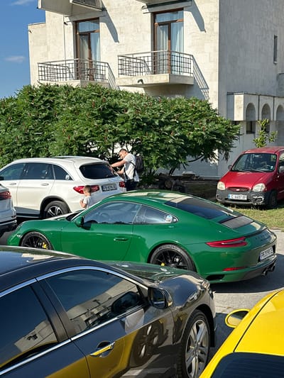The image depicts a parking lot in front of a building in Arbanassi, Bulgaria. A white SUV is parked next to a vibrant green Porsche. A man in a white t-shirt and black shorts, wearing a backpack, is interacting with the rear of the white SUV, possibly loading or unloading items. A young child in a white t-shirt is standing beside the rear of the white SUV, looking towards the man. Further to the right, a small red car is parked, showing some wear and tear on its front bumper. The building in the background appears to be a light-colored, multi-story structure with balconies and arched architectural elements. Lush green foliage, likely a dense shrub or tree, partially obscures the lower portion of the building. The ground is paved in the foreground, transitioning to a grassy area where the red car is parked. The scene is illuminated by bright sunlight, casting shadows on the building and the ground. The license plate on the white SUV reads "PB 4096 XE". The license plate on the green Porsche reads "PB 2090 CE". The license plate on the red car reads "C 6687 XP".