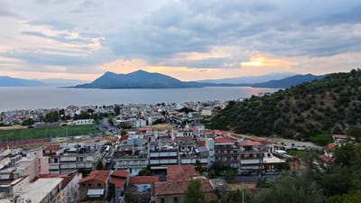 This is a wide-angle, outdoor, panoramic photograph of a coastal town in Greece, likely taken during dusk. The scene overlooks a body of water, possibly a bay or sea, with a prominent mountain range in the distance. The sky is filled with dramatic, heavy clouds, with patches of soft, warm light breaking through, indicating the setting sun. The light reflects on the calm water, creating a serene atmosphere.
The town itself is densely packed with buildings, characterized by white or light-colored facades and numerous red-tiled roofs, typical of Mediterranean architecture. There are several multi-story apartment buildings, some with balconies, and smaller residential homes. On the left side of the image, a green sports field, possibly a soccer pitch with a running track around it, is visible. Roads wind through the town, with a few cars parked along them.
The foreground is dominated by the town's buildings, seen from an elevated vantage point. The right side of the image features a lush, green hillside covered with olive trees and other vegetation, suggesting a rural or semi-rural setting encroaching on the urban landscape.
No people are distinctly visible, but the presence of the town and the sports field implies human habitation and activity. The overall mood is peaceful and picturesque, capturing the tranquil beauty of a Greek coastal town at sunset. The location is identified as Λουτρά Αιδηψού, Greece. No visible text is present in the image.