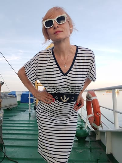 A woman with short, light-colored hair and white cat-eye sunglasses stands on the deck of a ferry. She is wearing a striped V-neck dress with a black belt featuring an anchor emblem. Her hands are on her hips. A life preserver hangs on a railing to her right. The deck is painted green, and in the background, the sky is overcast, and there are distant ships on the water. The location is the ferry terminal in Shchetinino, Russia.