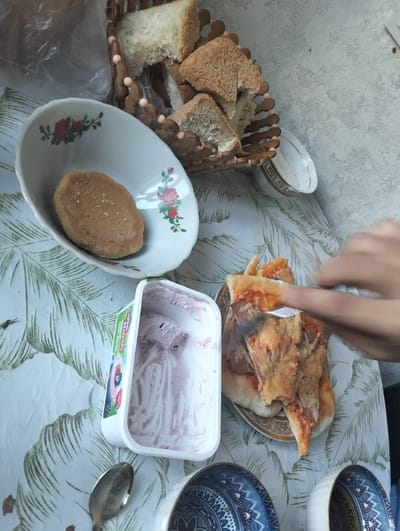 The image depicts a close-up, overhead view of a meal laid out on a table covered with a tablecloth featuring a tropical leaf pattern. The setting appears to be indoors, possibly a casual dining or picnic environment.
The primary subjects are food items and serving dishes. In the foreground, a partially eaten pizza is visible on a decorative plate. A hand is seen holding a fork, in the process of taking a slice of pizza. To the left of the pizza is a white container with a partially consumed purple-colored ice cream or dessert. Next to the ice cream container is a white oval-shaped dish containing a single, light brown, oval-shaped baked good, possibly a biscuit or bread roll, sprinkled with fine grains. Behind the dish, a wooden basket holds several pieces of torn bread. Further in the background, a small, white cup with a gold rim is visible.
The scene suggests a meal is in progress, with the pizza actively being served. The overall impression is one of informal enjoyment of food. There is no clear indication of the time of day or weather conditions. The location context is Aksu, Kazakhstan.
Visible text includes Russian on the ice cream container: "НОРМАЛ" and "ЖИДКОСТЬ" and "ДЕСЕРТ МАССА".