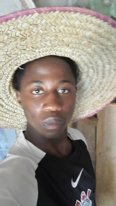 The image shows a close-up, head-and-shoulders shot of a young person wearing a straw hat. The hat is woven from light-colored straw with a pink band around the brim. The person's face is turned towards the camera, with their eyes open and looking forward. Their lips are slightly parted. The person is wearing a black t-shirt with a white Nike logo and some other graphics on the left chest area. The background is out of focus, but appears to be an indoor setting with a light-colored wall and some wooden elements. The lighting suggests it could be daytime. The image is taken from a slightly high angle, looking down at the subject. The setting is identified as Kabwir, Nigeria.