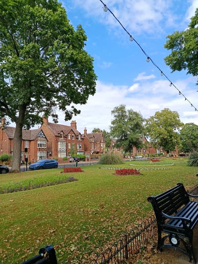 This image depicts a tranquil park scene with a row of traditional brick houses in the background. The park is characterized by lush green grass, scattered fallen leaves, and flower beds containing red and purple blooms. A black park bench is prominently featured in the foreground, overlooking the park. Trees with full green canopies stand tall, some framing the houses and others extending into the sky. A string of decorative lights hangs above the scene, suggesting it could be an evening setting or a place prepared for an event.
The houses are built in a classic architectural style, with multi-paned windows, pitched roofs, and brick facades. Some of the houses have decorative black and white timber framing on their upper levels. A blue hatchback car is parked in front of one of the houses, alongside a street lamp. The sky above is a clear blue with scattered white clouds, indicating pleasant weather. The presence of fallen leaves suggests it might be autumn.
No people are visible in the image. The overall mood conveyed is one of peacefulness and quietude, with the well-maintained park and classic architecture creating a sense of established charm. The location is identified as Bedford, United Kingdom. There is a small circular logo visible on the bench, but the text within it is not legible.
