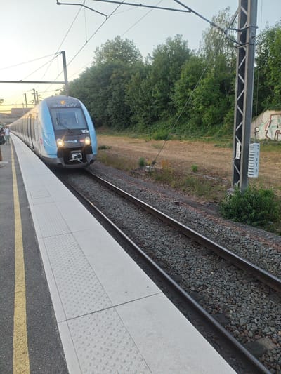 A bright blue train with a black front and yellow accents is stopped at a station platform in Vaires-sur-Marne, France. The train has "RÉSEAU FRANCE" and "SNCF" branding visible on its front. A person in a white shirt and dark shorts is standing on the platform to the left of the train. The platform is paved with concrete tiles that have raised circular patterns, and a yellow line is visible along the edge of the asphalt path. Two sets of train tracks run alongside the platform, filled with gray gravel. A tall metal utility pole with electrical wires attached stands near the tracks, with graffiti visible on a wall behind it. Trees and dry grass line the embankment behind the tracks. The sky is clear and light, suggesting either early morning or late afternoon.