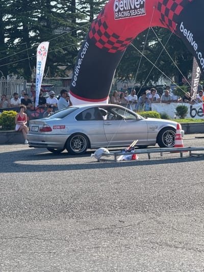 In Poti, Georgia, a silver BMW E46 coupe is positioned on an asphalt surface, partially obscured by a red and black checkered archway. The archway is branded with "betlive.com RACING SEASON." A crowd of spectators is visible behind a railing. To the left, a woman in a red and white outfit is seated on a low wall. The car displays a "GDS PARK DRIFT" sticker on its rear. Orange and white traffic cones and flags are present near the car. The scene appears to be an outdoor event, likely a race or car show, under daylight.
