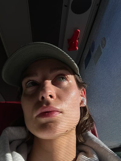 A close-up, low-angle, natural light shot features a young person wearing a green baseball cap and a grey hooded sweatshirt. The person's face is illuminated by a striped pattern of light and shadow, suggesting they are near a window with blinds. They are looking upwards, with their eyes open and focused. In the background, a red object, possibly a hook or a handle, is attached to a dark ceiling. The scene appears to be indoors, possibly on a bus or train, given the window and overhead features. The location is not specified as Neumünster, Germany.