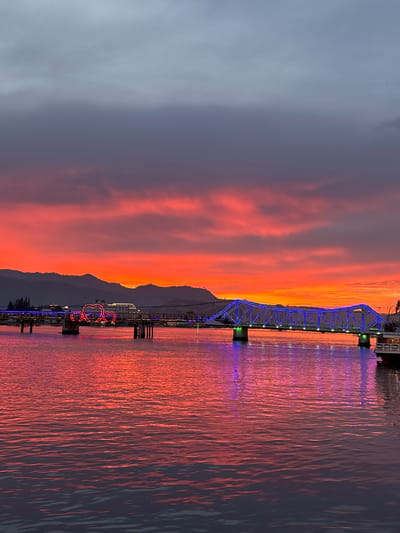This is a photograph of the Kampot River in Kampot, Cambodia, at sunset. The sky is a dramatic gradient of deep purple, vibrant orange, and bright red, with dark clouds overhead. The water of the river reflects the colors of the sunset, creating a shimmering, pinkish-red surface.

A bridge spans the river, decorated with bright blue lights along its structure. On the left side of the bridge, there are three illuminated heart-shaped structures, also in bright pink neon. The bridge is supported by concrete pillars. In the background, silhouetted mountains rise against the horizon. On the far right, a portion of a white boat with a dark hull is visible docked at the river's edge. There are no people visible in the photograph. The scene is illuminated by the natural light of the sunset and the artificial lights on the bridge and heart structures.