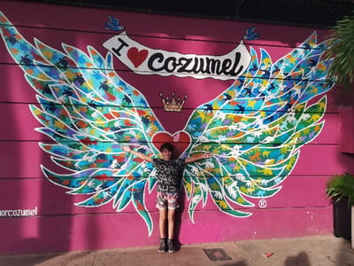 The image shows a mural on a pink wall in Cozumel, Mexico. The mural depicts two large wings, made up of many colorful, stylized sea turtles, seabirds, and fish. The mural is located in the downtown area of Cozumel (latitude: 20.511, longitude: -86.95). A young boy wearing a black t-shirt, blue shorts, and black shoes is standing in front of the mural, with his arms outstretched. He is touching the wings. There is a sign above the wings that says "I love Cozumel." The sign is in the shape of a heart with a white outline and is decorated with a small red heart. The mural is located on the wall of a building with a pink facade, and the wall is surrounded by greenery and palm trees. The mural is likely located in the city's tourist district.