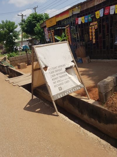 The image depicts an outdoor scene in Palade, Nigeria, likely a street or marketplace. The main subject is a tilted, white signboard mounted on a wooden frame, displaying services offered. The text on the signboard indicates "ACCOUNT OPENING," "CASH DEPOSIT," "CASH WITHDRAWAL," "MONEY TRANSFER," "PAY BIELS (DSTV, GOTV & STARTIMES)," and "RECHARGE CARDS." A phone number, "08141930204," is also visible for inquiries.

Behind the signboard, shelves stocked with various products, including Maggi, Nestlé, Nido, and possibly toiletries, suggest a small shop or kiosk. An MTN branding for "MoMo Airtime Data Broadband" is also visible, indicating a telecommunication service provider. A broom made of dried grass stands nearby, a common sight in many African settings. The ground is a mix of compacted earth and gravel, with a concrete drainage channel running alongside it.

The scene appears to be set during the daytime, with clear shadows indicating sunlight. The presence of trees and a glimpse of a blue sky suggest a warm, possibly humid climate. The overall impression is one of a bustling local commercial area, with services catering to everyday needs and telecommunication demands. There are no people visible in the frame, but the setup implies an active business. The image has a slightly tilted perspective, possibly due to the camera angle.