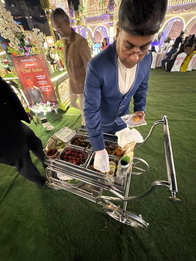 A man in a blue jacket and white gloves serves food from a silver cart with multiple compartments holding various items like olives and fruit. The cart is on green turf, and a sign displays "Chef Goku's" name and image. A man in a tan jacket stands nearby, and people sit at decorated tables in the background. The event appears festive with floral arrangements and ornate architectural details. The focus is on the food service and branding of the chef.