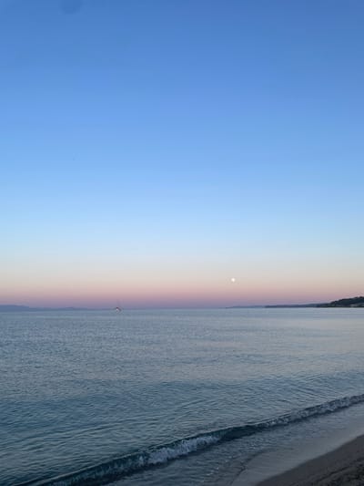 A wide-angle, eye-level shot captures a serene seascape at dusk in Nea Potidea, Greece. The sky transitions from a deep, clear blue at the top to soft hues of pink and lavender near the horizon, indicating the setting sun. A small, white sailboat is visible in the middle distance, sailing on the calm, blue-green water. A full moon is a pale disc in the sky to the right of the sailboat. The shoreline, composed of sand and small pebbles, occupies the bottom right portion of the frame, with gentle waves lapping at the beach. In the far distance, a faint outline of land, possibly a coastline or mountains, is visible against the colorful sky. The scene is devoid of any people or visible activity, emphasizing the tranquility of the coastal environment.