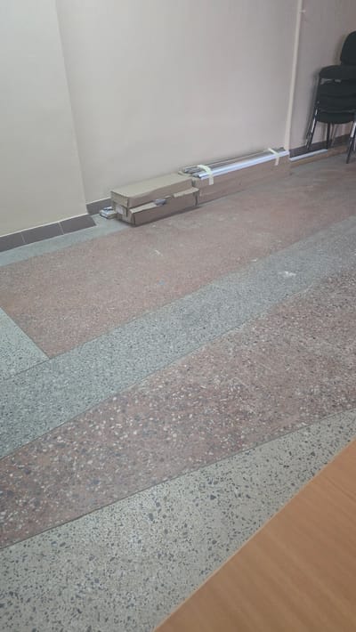 The image shows a hallway with a light beige wall and a terrazzo floor. Two cardboard boxes are stacked against the wall, with one on top of the other. The top box is partially open and appears to contain metallic rods. The bottom box is closed and taped shut. There are also some smaller packages or boxes stacked next to the larger ones. In the background, there are a few black chairs stacked on top of each other. The overall scene suggests a storage area or a delivery point within an indoor environment. The location of Ukhta, Russia, is not visually discernible from the image itself.