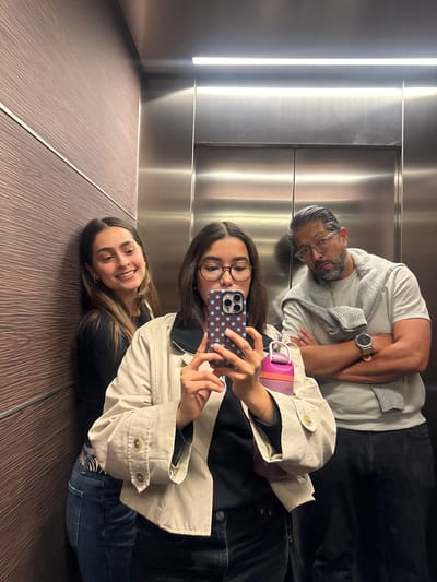 Three individuals are captured in an elevator interior. One young woman with long brown hair and wearing a black top and blue jeans is smiling while laying on the wooden patterned floor of the elevator. Above her, a second young woman with glasses is holding a mobile phone with a blue polka dot case, seemingly taking a picture of the group. She is also wearing a jacket. Above them and to the side is a man with salt and pepper hair and a beard, wearing a light-colored shirt and a sweater. The walls of the elevator are stainless steel. The location is not evident in the image and cannot be determined to be in Santiago Yancuitlalpan, Mexico.