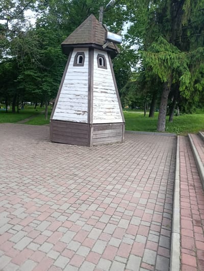 This is a medium-angle, eye-level shot of a decorative windmill-like structure in a park setting. The structure is made of wood and painted white with brown trim. It has a conical roof and two arched windows on its visible side. The base of the structure is dark brown, also made of wood.
The windmill is situated on a paved walkway made of interlocking red and grey bricks. To the right of the walkway, there are concrete steps also lined with similar bricks. In the background, there are numerous mature trees with lush green foliage, suggesting a temperate climate and possibly late spring or summer. The ground in the park area is covered in grass.
There are no people clearly visible in the immediate vicinity of the windmill, though there appear to be some blurred figures in the distance on the grass. The lighting suggests it is daytime, and the overcast sky implies a cloudy or diffused light condition.
The overall scene is peaceful and appears to be a public park or recreational area in Khorol, Ukraine. The windmill serves as an ornamental feature. There is no visible text in the image.
