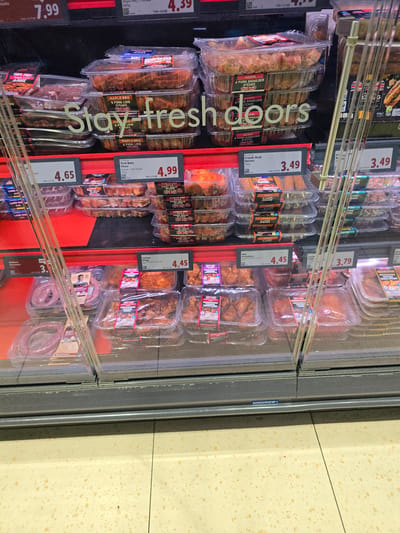 An interior, eye-level, medium shot captures a refrigerated display case filled with various cuts of meat products. The display case features multiple shelves stacked with plastic-wrapped meat packages, primarily pork and lamb products, with some chicken drumsticks visible. Several price tags are affixed to the front of the shelves, displaying prices such as £4.65, £4.99, £3.49, £4.45, and £3.79. A large, faded white text overlaid on the image reads "Stay fresh doors". The background is dark, with vertical metal support beams of the display case visible. The floor is tiled with a cream-colored speckled pattern. The lighting suggests an indoor retail environment, possibly a supermarket or butcher shop. The overall impression is one of a well-stocked meat counter. The image is set in Stockton-on-Tees, United Kingdom.