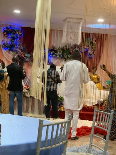 A wedding scene includes floral decorations and beaded curtains. Several people are standing around. One man wears a white outfit with matching pants. Another man is in a plaid shirt with jeans, and a third man wears a black jacket. Chairs with a blue tablecloth can also be seen. The photo appears to be taken indoors, possibly at a reception hall.