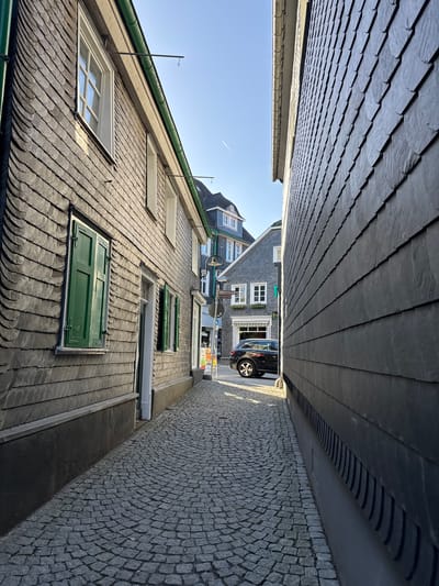 The image shows a narrow alleyway in Remscheid, Germany. The alley is flanked by buildings with slate-tiled walls and roofs. One building on the left has green shutters and white-framed windows. The alley is paved with cobblestones. A black car is parked further down the alley. The sky is visible at the end of the alley. There are no people visible in the image.