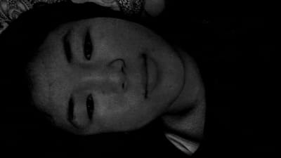 The image shows a black and white, close-up, overhead shot of a person's face. The person appears to be lying down, with their head tilted. The lighting is dim, casting shadows that obscure some of the facial features. The background is dark, with a hint of a patterned fabric visible at the top left of the image. There are no discernible landmarks or details that would indicate a specific location within the city of Uchkun, Kyrgyzstan. The image focuses solely on the subject's face and their current resting state.
