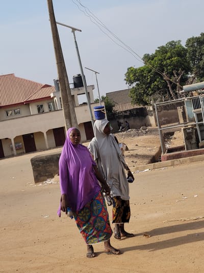 The image depicts a daytime street scene in Nassarawa Egon, Nigeria. Two adult females are walking from the right towards the left across a dusty, unpaved road in the foreground.

The first female, on the left, wears a solid purple head covering (hijab) and a long, patterned skirt featuring blue, red, and yellow geometric and floral motifs. She carries a small blue plastic bottle in her right hand and wears dark sandals.

The second female, positioned slightly behind and to the right of the first, wears a light grey head covering and a loose, long-sleeved grey garment. She carries a blue cylindrical container balanced on her head and a blue plastic bottle in her left hand. She wears dark sandals, and gold-colored wrist jewelry is visible on her right arm.

In the mid-ground, on the left, stands a two-story building with light-colored walls and a terracotta-colored pitched roof. The ground floor features multiple arched openings, some with closed brown doors. Above this building, a tall concrete utility pole with overhead wires is visible. Adjacent to the building on the right, another structure supports two dark water tanks on a raised platform. To the right of the concrete pole, a thinner metal pole with a solar street light fixture is present.

Further in the background, on the right side of the road, a concrete wall separates the street from a dirt area containing refuse. Two individuals, appearing to be males, are visible sitting or crouching near this wall amidst the refuse. To the far right, a metal cage structure, possibly an enclosure for utilities, contains debris. Lush green trees are visible beyond the wall and structures in the background. The sky is clear and light blue, indicating bright daylight with strong shadows cast on the ground. The road surface contains scattered paper and debris.