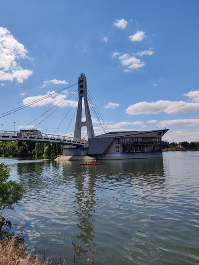 A wide shot captures a modern suspension bridge and a multi-story building complex on a body of water on a clear, sunny day. The bridge features a distinctive tall, angled concrete tower with cables extending outwards to support the walkway. To the right of the tower, a contemporary, angular building with a dark roof and numerous windows is situated on a platform over the water. In the foreground, a small red kayak with several people wearing life jackets is paddling on the rippling water. The water's surface reflects the sky and the surrounding structures. The background shows lush green trees along the riverbank and a few houses in the distance. The sky is a bright blue with scattered white clouds. The image is taken from a slightly elevated viewpoint, looking across the water towards the bridge and the building. The location is identified as Novaya Adygeya, Russia.