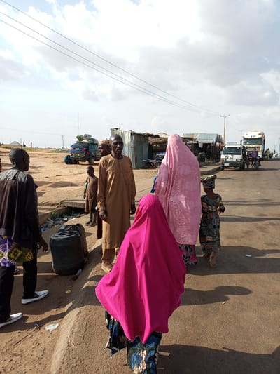 In Gadao, Nigeria, a group of people are gathered near a road. A man in a brown traditional robe stands on the right, facing the viewer, with a young boy in a similar robe beside him. To the left, two individuals are seen from the back, one wearing a brown jacket and patterned fabric, the other partially obscured. In the foreground, a person in a bright pink headscarf is facing away from the camera. Further in the background, two women in traditional attire are walking away, one in a pink head covering and the other in a patterned dress and headscarf. A small child in a colorful dress walks alongside them.

The environment is dusty, with a dirt path running alongside the paved road. Several vehicles are visible, including a dark blue SUV with luggage on top, a white truck, and a smaller white vehicle. In the middle ground, there are makeshift structures with corrugated metal roofs, suggesting a market or roadside stalls. Large black plastic containers are placed on the side of the path. Overhead, power lines stretch across the sky, which is partly cloudy. The scene appears to be an outdoor gathering or transit point.
