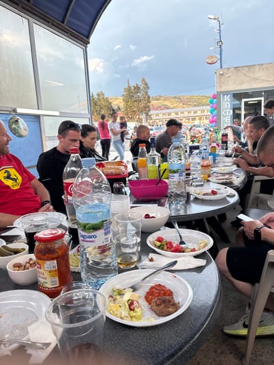 A photograph captures a table laden with various food and drink items, with people seated around it, presumably dining together. The table holds plates containing salads and meat, an assortment of bottled water brands, fruit juice, and condiments in jars. There are also plastic cups and bowls containing nuts and pickles.

The setting appears to be an outdoor dining area. In the background, across a street, there is a building adorned with a mural or a large advertisement featuring balloons and a mountainous landscape. The signage visible suggests a location in Aytos, Bulgaria. Individuals can be seen both at the table and in the background, possibly pedestrians or other patrons.