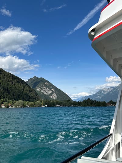 The image captures a view from a white boat with a red stripe, sailing on a clear blue lake under a bright blue sky with scattered white clouds. The boat is angled to the right of the frame, showing its railing and side. In the background, a lush green mountainside with dense trees rises from the opposite shore, dotted with houses. Further in the distance, more majestic mountains with rocky grey peaks are visible. The water is a vibrant turquoise, with visible ripples and waves created by the boat's movement. The scene suggests a scenic journey through the Swiss landscape, likely on Lake Thun, near Sigriswil, Switzerland. There are no people visible in the image.