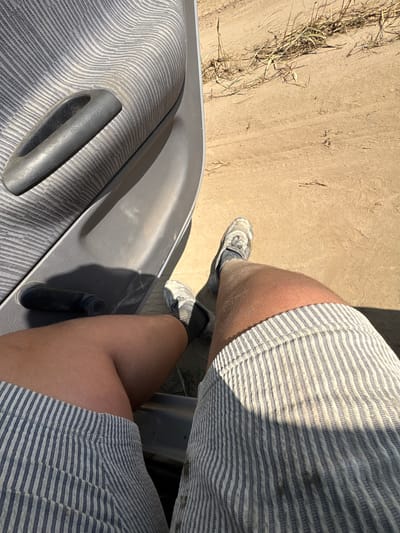 The image shows a person's legs and a portion of a car's interior, viewed from a first-person perspective. The person is wearing striped shorts and dirty sneakers. The car interior includes a grey fabric door panel with a dark grey handle and a black window crank. The exterior scene visible through the open car door is a dirt or gravel path with some dry grass and straw. The lighting suggests it is daytime. There is no identifiable information that places this scene specifically in Opatów, Poland.