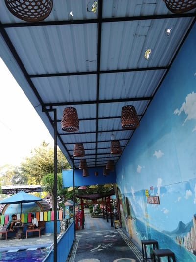 An outdoor walkway runs alongside a building with a blue mural depicting a seascape. The walkway is covered by a corrugated metal roof supported by a black metal frame. Several hanging basket-shaped lamps are suspended from the ceiling. On the left side of the image, two young males are seated at a table under a blue umbrella, near a swimming pool. Trees and foliage are visible in the background. The mural on the right wall features a body of water, mountains, and a cloudy sky, with a sign that reads "KEDALAMAN 1,5 M" (Depth 1.5 M). Two dark wooden stools are placed in front of the mural. The walkway itself is paved with dark asphalt decorated with colorful circular patterns.