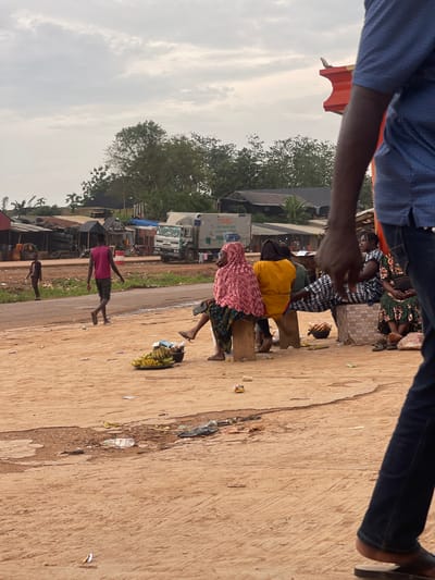 The image depicts a bustling market scene in Osu, Nigeria, likely in the afternoon given the warm lighting. The sky is overcast, suggesting a cloudy day. The foreground is dominated by a dusty, textured ground, littered with various pieces of trash, including plastic bottles and wrappers, indicative of a busy public space.
Several people are visible, engaged in activities typical of a market. In the middle ground, three women are seated on wooden benches. One woman, wearing a pink headscarf and patterned skirt, is looking down. Beside her, another woman in a yellow top is also seated. Further back, a woman in a black and white patterned dress is partially visible.
To the left of the seated women, a young person in a pink shirt and dark shorts stands, looking towards the left. Their posture suggests they might be observing something or in motion. Behind them, a chaotic collection of market stalls and vehicles can be seen, including what appears to be a white truck with some visible text: "CASE LTD". The overall scene conveys a sense of everyday life and commerce in a Nigerian community. The presence of multiple individuals and the market setting suggest it is a lively and active environment.