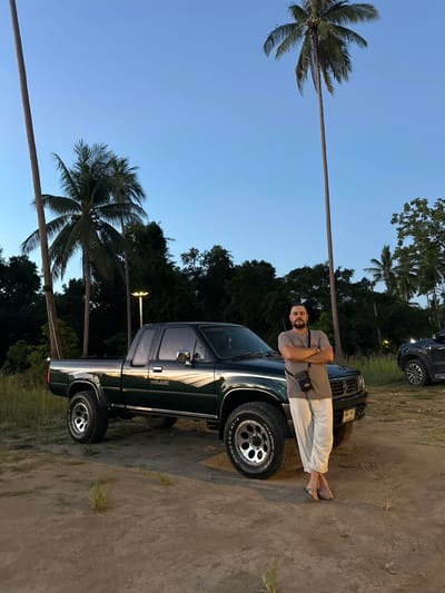 A man stands with his arms crossed in front of a dark green Toyota Hilux pickup truck. The man is wearing a grey t-shirt and light-colored pants, with sandals on his feet. He has a small crossbody bag. The truck is parked on a dirt lot with sparse grass. In the background, there are tall palm trees and dense foliage. The sky is a clear blue, indicating it is likely dusk or dawn. A street light is visible in the distance. To the right of the Hilux, part of a dark-colored SUV is visible. The scene is located in Baan Taling Ngam, Thailand, likely near a beach or coastal area, given the presence of palm trees.