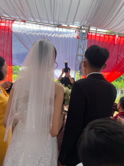 This image captures a moment at a wedding ceremony or reception in Tay Son Commune, Vietnam, observed from behind the bride and groom. The setting is under a decorated tent or canopy.
The main subjects are the groom, seen from the back and left side, wearing a black suit jacket over a white collared shirt, and the bride, positioned to his right, also viewed from behind. The bride is adorned in a white, off-the-shoulder wedding dress featuring lace detailing. A long, sheer white veil, embellished with small pearl-like beads, cascades over her back. She holds a white floral bouquet, partially visible between her arm and the groom.
In the background, a person, likely a photographer, is visible holding a black camera with an attached flash unit, actively capturing the event. Other attendees are partially obscured around the edges of the frame.
The event takes place within a structure made of metal trussing, draped with panels of white and vibrant red fabric, indicating a festive occasion. Bright natural light filters through the canopy, suggesting it is daytime. Green foliage is visible in the distance beyond the canopy, indicating an outdoor or semi-outdoor location. No visible text is present in the image.
