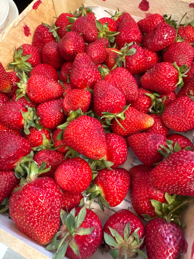 A close-up shot displays a wooden crate brimming with ripe strawberries. The strawberries are predominantly red, with green stems and leaves. Some appear shiny, possibly due to moisture. The background of the crate shows faint Russian script. The setting might be a local market or farm in Тоншалово, Russia, given the likely origin of the packaging. No people are visible.