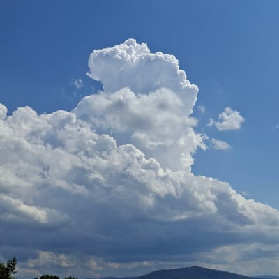 This is a photograph of a sky filled with dramatic, towering cumulonimbus clouds. The clouds are white and puffy, with darker, shadowed undersides suggesting their immense size and vertical development. The sky is a vibrant blue, indicating it is daytime, likely late morning or early afternoon given the bright sunlight illuminating the clouds.
The image appears to be taken from ground level, looking upwards. The perspective is vertical, as evidenced by the orientation of the clouds and the background elements. On the right side of the frame, a steep, tree-covered mountainside is visible in silhouette, partially obscured by a large cloud formation. Power lines stretch vertically along the mountainside, suggesting proximity to a populated or developed area. The presence of a forest on the mountainside and the clear blue sky are typical of a fair weather day, though the large cumulonimbus clouds hint at potential for developing storms.
The overall mood of the image is one of awe and natural grandeur. The sheer scale of the clouds is the dominant feature, creating a sense of powerful natural forces at play. There are no people or man-made objects that are the primary subject, only the vast expanse of sky and cloud with subtle hints of the landscape below. The location context of Baden-Baden, Germany, suggests a mountainous and perhaps picturesque European setting. There is no visible text in the image.