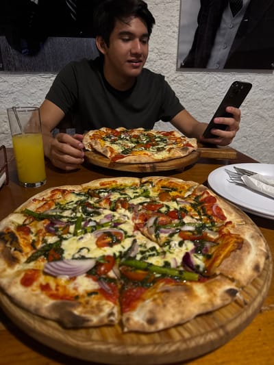 A young man in a dark green t-shirt is photographing two pizzas with his smartphone. The pizzas are on wooden serving platters. One pizza is smaller and appears to have a simple cheese and tomato topping. The larger pizza has toppings including tomatoes, red onion, spinach, and asparagus. A plate of utensils in a napkin is on the table between the pizzas. A glass of yellow liquid is in the foreground. The background features a partially visible framed print and a textured wall. The location is likely a restaurant in Chipilo de Francisco Javier Mina (Chipilo), Mexico.