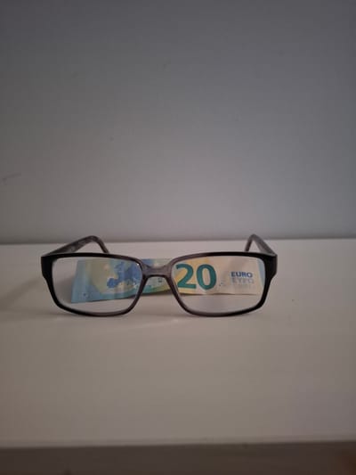 A pair of dark-rimmed eyeglasses is positioned horizontally on a light-colored surface, likely a table or shelf. Behind the eyeglasses, a portion of a 20 Euro banknote is visible. The banknote features a map of Europe and the number "20" prominently displayed. The background is a plain, light grey wall. The lighting is neutral, casting soft shadows. There is no indication of the image's location in Huntingdonshire, United Kingdom.