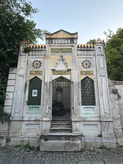 The image depicts the exterior of a tomb structure in Eyüpsultan, Turkey. The tomb is constructed from marble and features intricate golden Arabic calligraphy on its facade. A metal gate stands in front of the entrance, and a QR code is visible on one of the windows. The structure is flanked by trees, and the foreground is paved with cobblestones. The sky above is a clear, pale blue, suggesting it is either early morning or late afternoon. The overall scene conveys a sense of historical and religious significance.