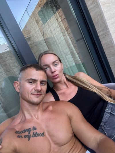The image shows a close-up, selfie-style shot of a man and a woman. The man is in the foreground, shirtless, with a tattoo on his chest that reads "boxing is in my blood". He is looking towards the camera with a slight smile. The woman is behind him, resting her head on his shoulder. She has long blonde hair and is wearing a black top and denim shorts. Both individuals appear to be tanned, suggesting a sunny environment.

The setting appears to be outdoors, possibly on a balcony or patio, with large glass doors reflecting a textured stone wall and a ribbed metal ceiling. The angle of the shot and the reflections suggest the image was taken during the daytime. The overall mood seems relaxed and intimate. The location context provided is Finestrat, Spain, which aligns with the sunny, outdoor setting.