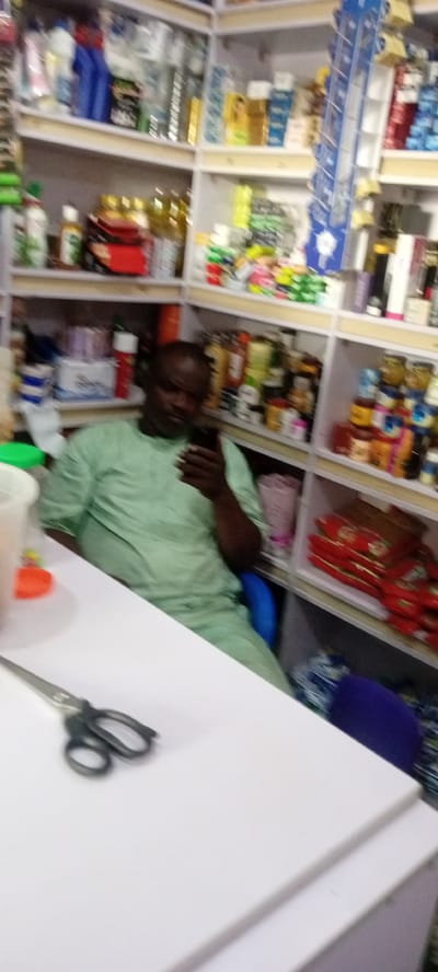 The image shows a man sitting in a shop in Gayawa, Nigeria, looking at his phone. He is wearing a light green traditional outfit. The shop shelves are stocked with various products, including cleaning supplies, food items, and personal care products. A pair of scissors is visible on the white table in the foreground. The overall impression is of a small business in a commercial setting.