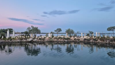 This image captures an artificial island constructed in the Mar Piccolo lagoon, a part of the Mar Grande, in Taranto, Italy. The island, adorned with numerous small white crosses, reminiscent of a cemetery, is situated against a twilight sky displaying hues of pink and blue. The crosses vary in size and design, some topped with decorative elements, and are interspersed with small trees, shrubs, and rocks. The water in the foreground reflects the island and the sky, creating a mirror-like effect. In the background, the mainland of Taranto is visible with a line of trees and buildings. The scene suggests a serene, somewhat melancholic, and artistic arrangement within the natural landscape of the lagoon.