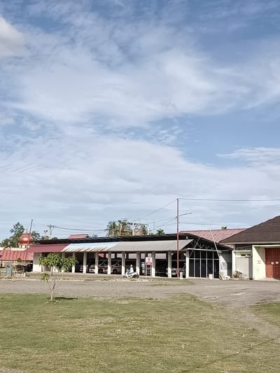 The image captures a scene in front of a building with a covered walkway, possibly a marketplace or public facility, with a dome-shaped structure visible to the left. The building has a long, open-sided roof supported by pillars, with a colorful section in the middle featuring blue and red panels. Several vehicles, appearing to be ambulances, are parked under the covered area. A person is riding a scooter in front of the building.
The foreground is dominated by a well-maintained green lawn with a small, sparse tree in the center. Beyond the lawn, there's a gravel area leading up to the building. The sky above is a clear blue with scattered, wispy white clouds, suggesting it is daytime, likely late morning or early afternoon given the lighting. The overall atmosphere appears calm and peaceful.
In the background, more buildings with pitched roofs are visible, along with trees and utility poles. The dome-shaped structure on the left, with its reddish-gold hue, suggests a religious building, possibly a mosque, given the location context of Mesjid Reuntoh, Indonesia. The architecture of the main building, with its open design and corrugated roofing, points to a tropical or subtropical climate.
There is a visible "P" sign with a circle around it, indicating a parking area. The time of day is suggested by the bright daylight and shadows cast. The weather appears to be pleasant and sunny. The image does not convey any strong emotions, presenting a factual depiction of the location.