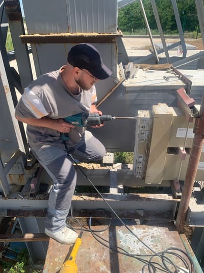In Ivanivka, Ukraine, a man is performing a task that involves drilling into a concrete structure. He is wearing a grey t-shirt with white and orange stripes on the sleeves, grey work pants, a black baseball cap, and glasses. He is holding a blue and black rotary hammer drill with both hands and appears to be focused on his work. The drill is actively engaged in the concrete, with dust and debris falling from the point of contact.

The setting is an outdoor industrial or construction site, with visible metal scaffolding and structures. A large, light-colored concrete block is being worked on, suspended by metal supports. There are electrical wires and tools, including a yellow power tool that resembles an angle grinder, scattered around the scene. The ground appears to be dirt or gravel, with some dry grass and vegetation visible in the background. The lighting suggests it is daytime, likely mid-morning or afternoon, under clear or partly cloudy skies. The overall mood is one of focused labor. One of the concrete blocks has a label on it, but the text is not clearly legible. The scene appears to be well-lit, indicating a bright day.