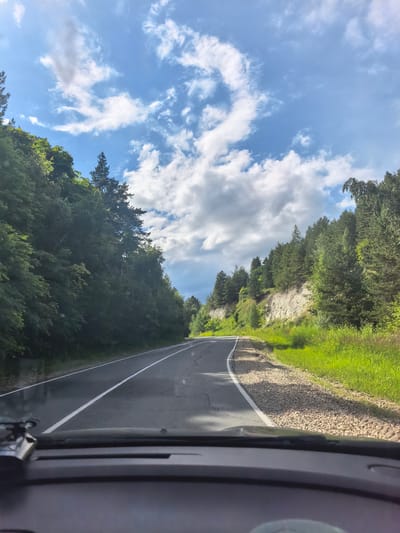 The image shows a view from the driver's seat of a vehicle traveling on a paved road. The road is bordered by lush green trees on the left and a rocky incline with trees on the right. The sky above is a vibrant blue with scattered white clouds. The road appears to be a two-lane highway with white lane markings. The terrain suggests a natural, possibly rural or mountainous environment. No people or specific landmarks are visible. The overall impression is that of a scenic drive through a natural landscape.